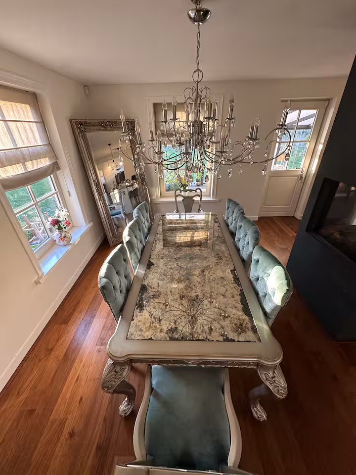 Formal dining table with tufted upholstered chairs under a silver chandelier, beside a large ornate mirror