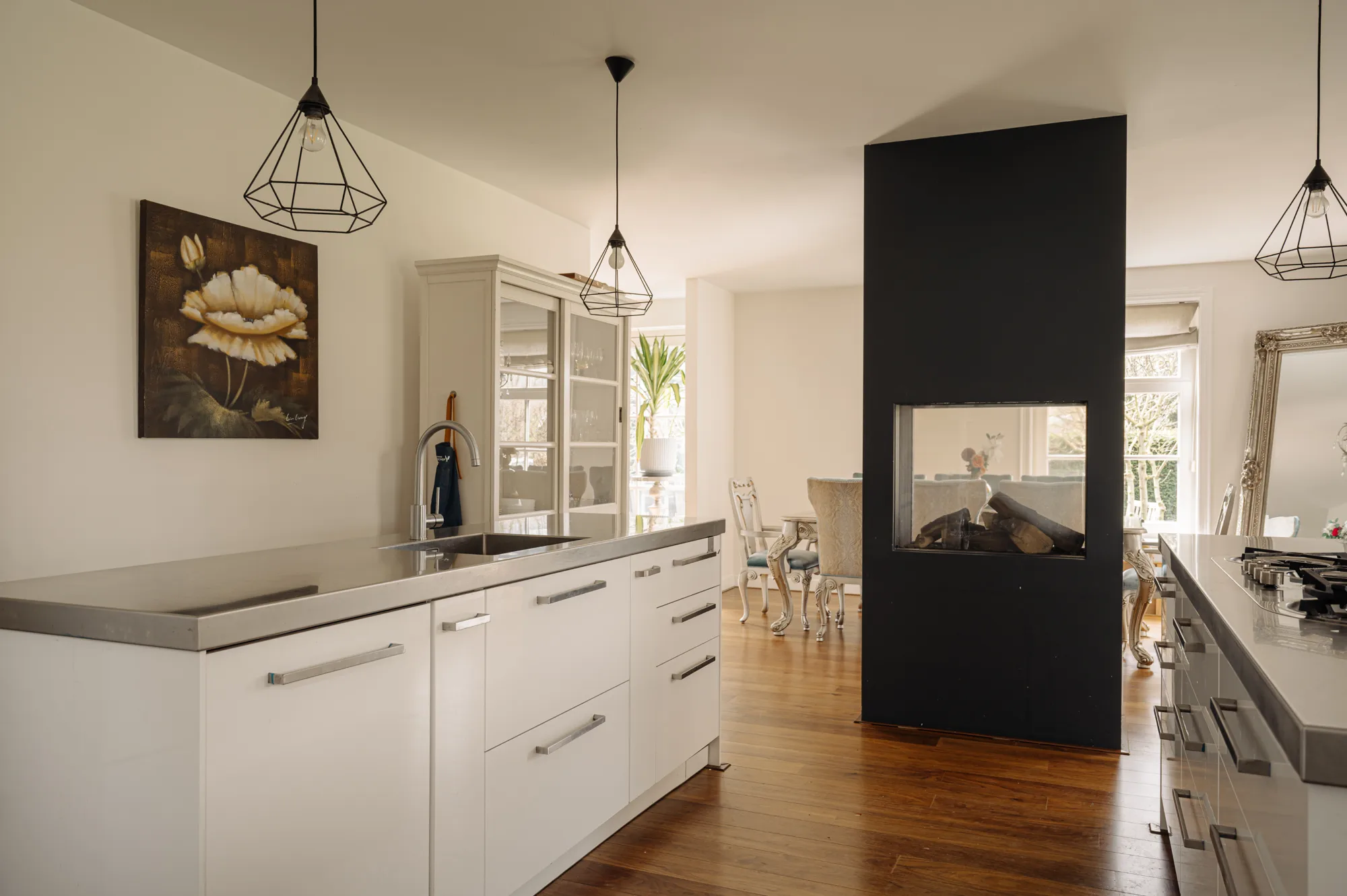 Kitchen island with a sink and gas cooktop beside a double-sided fireplace, with a view into the dining area